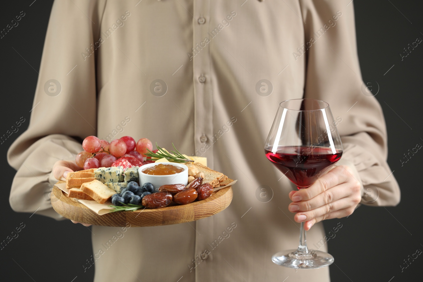 Photo of Woman holding board with different types of delicious cheese, other snacks and wine on grey background, closeup