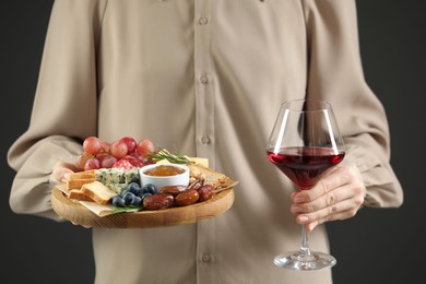 Photo of Woman holding board with different types of delicious cheese, other snacks and wine on grey background, closeup