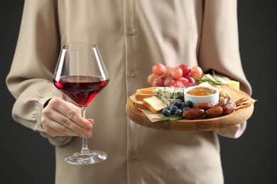 Photo of Woman holding board with different types of delicious cheese, other snacks and wine on grey background, closeup