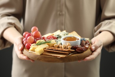 Photo of Woman holding board with different types of delicious cheese and other snacks on grey background, closeup