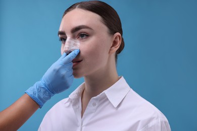 Doctor checking patient's nose after plastic surgery operation on light blue background, closeup