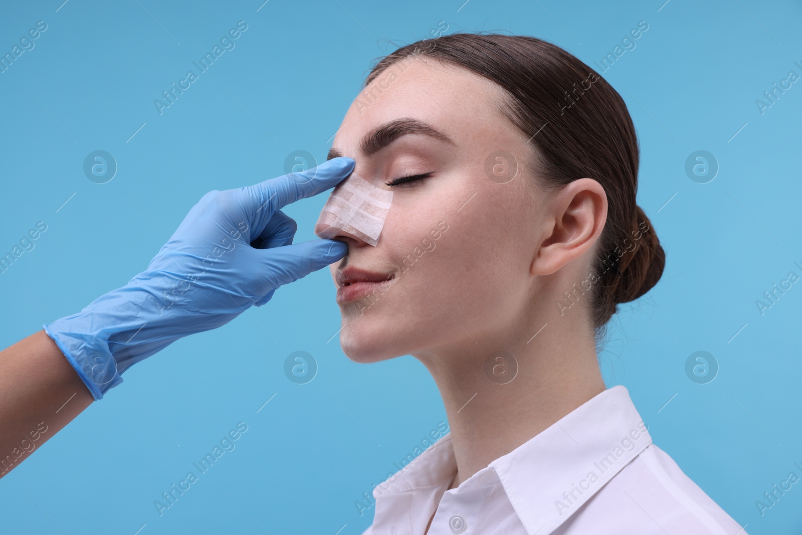 Photo of Doctor checking patient's nose after plastic surgery operation on light blue background, closeup