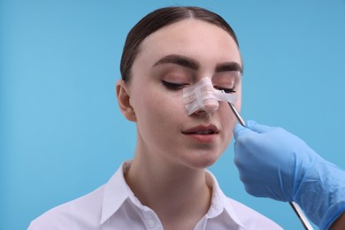 Doctor removing medical bandage from patient's nose after plastic surgery operation on light blue background, closeup