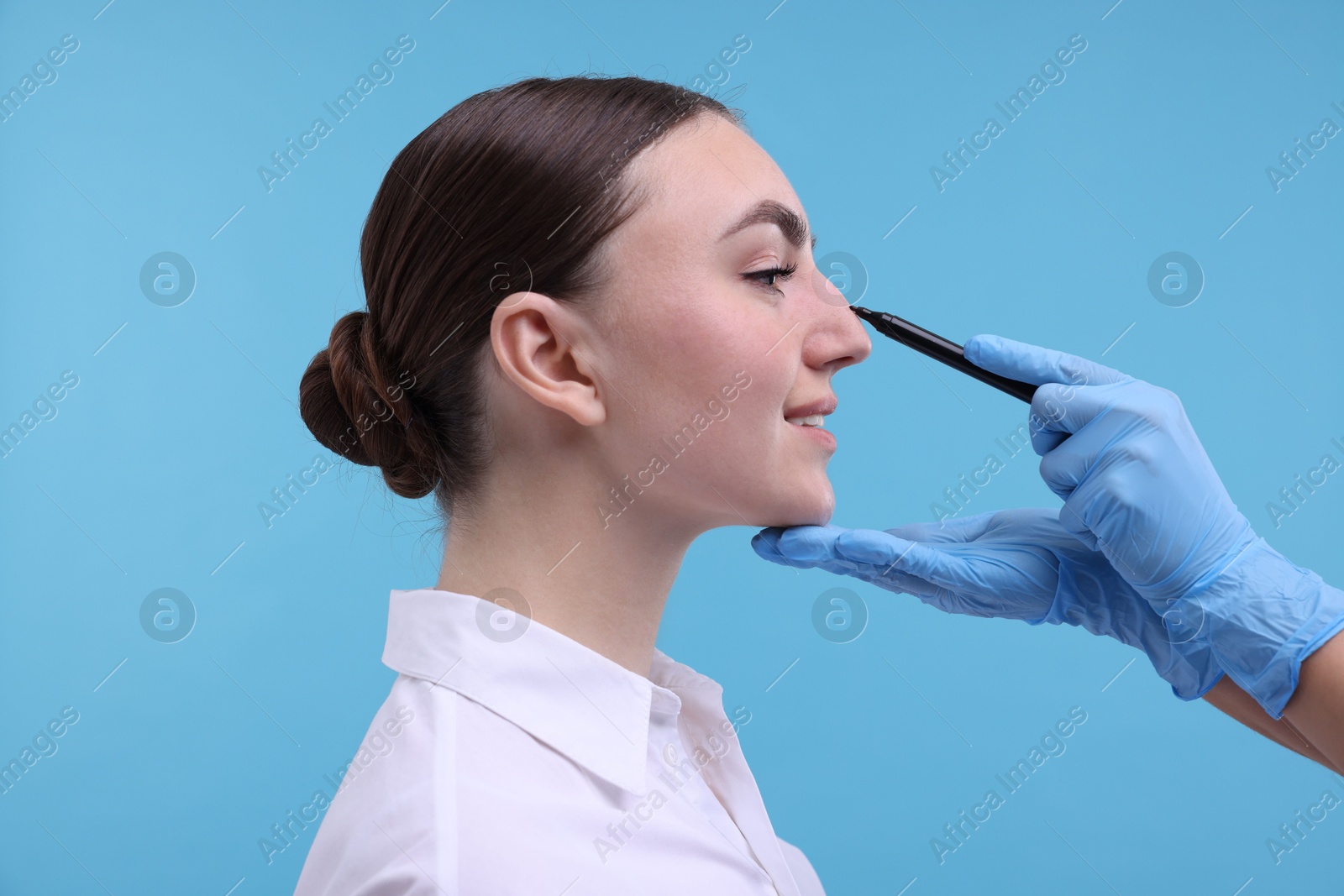 Photo of Doctor drawing marks on patient's nose for plastic surgery operation against light blue background, closeup