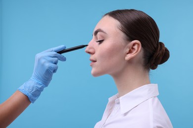 Doctor drawing marks on patient's nose for plastic surgery operation against light blue background, closeup