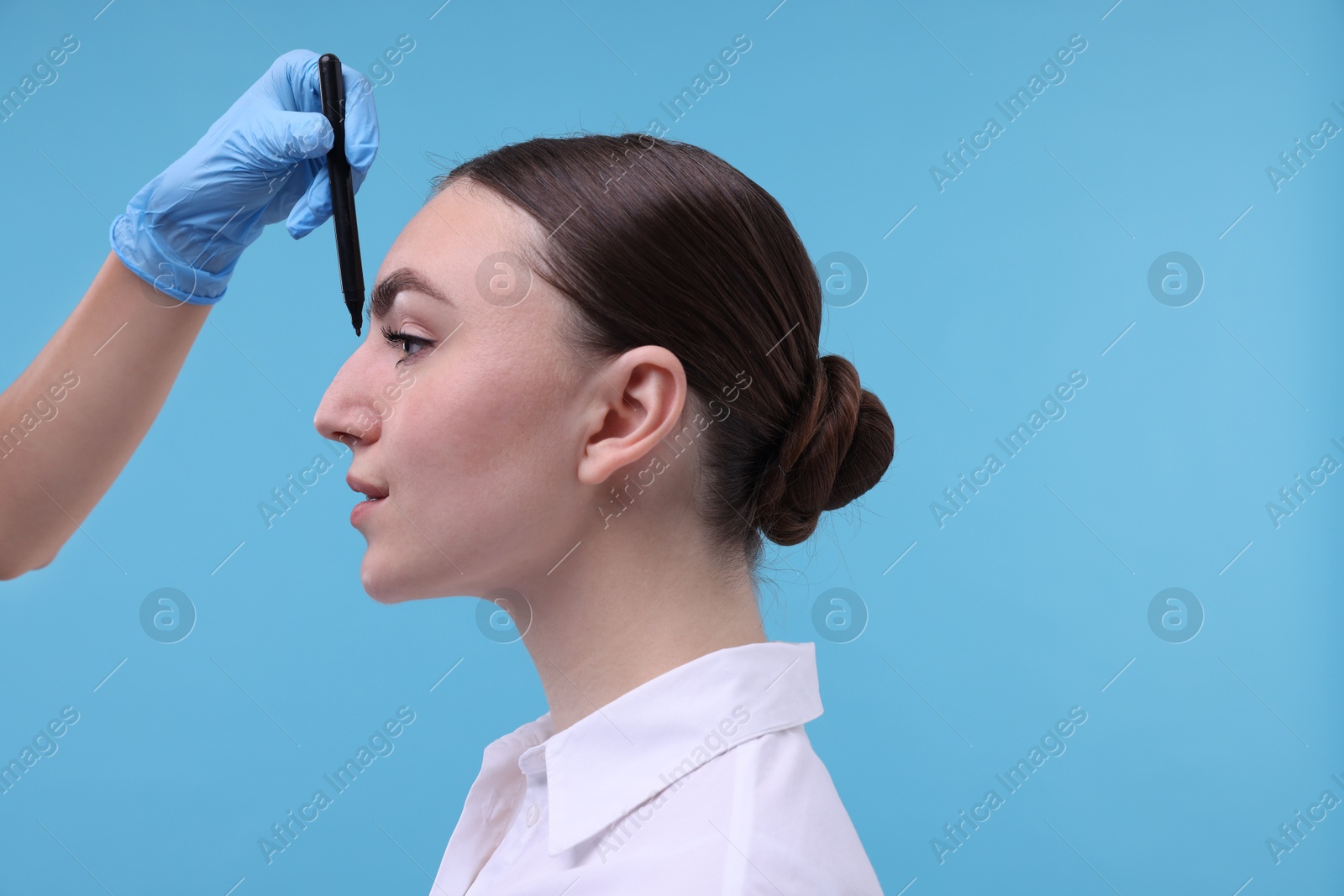 Photo of Doctor drawing marks on patient's nose for plastic surgery operation against light blue background, closeup