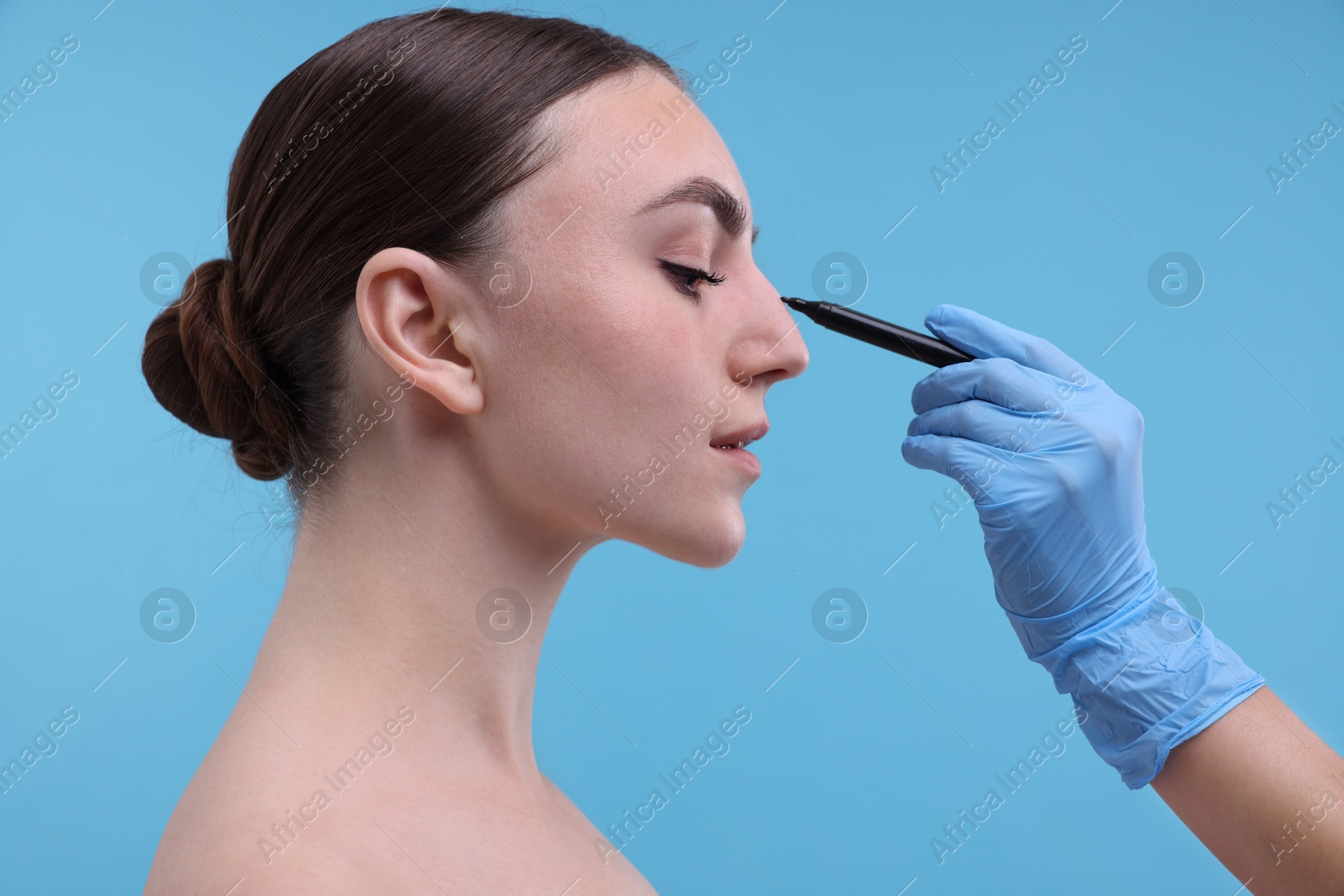 Photo of Doctor drawing marks on patient's nose for plastic surgery operation against light blue background, closeup