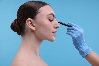 Doctor drawing marks on patient's nose for plastic surgery operation against light blue background, closeup