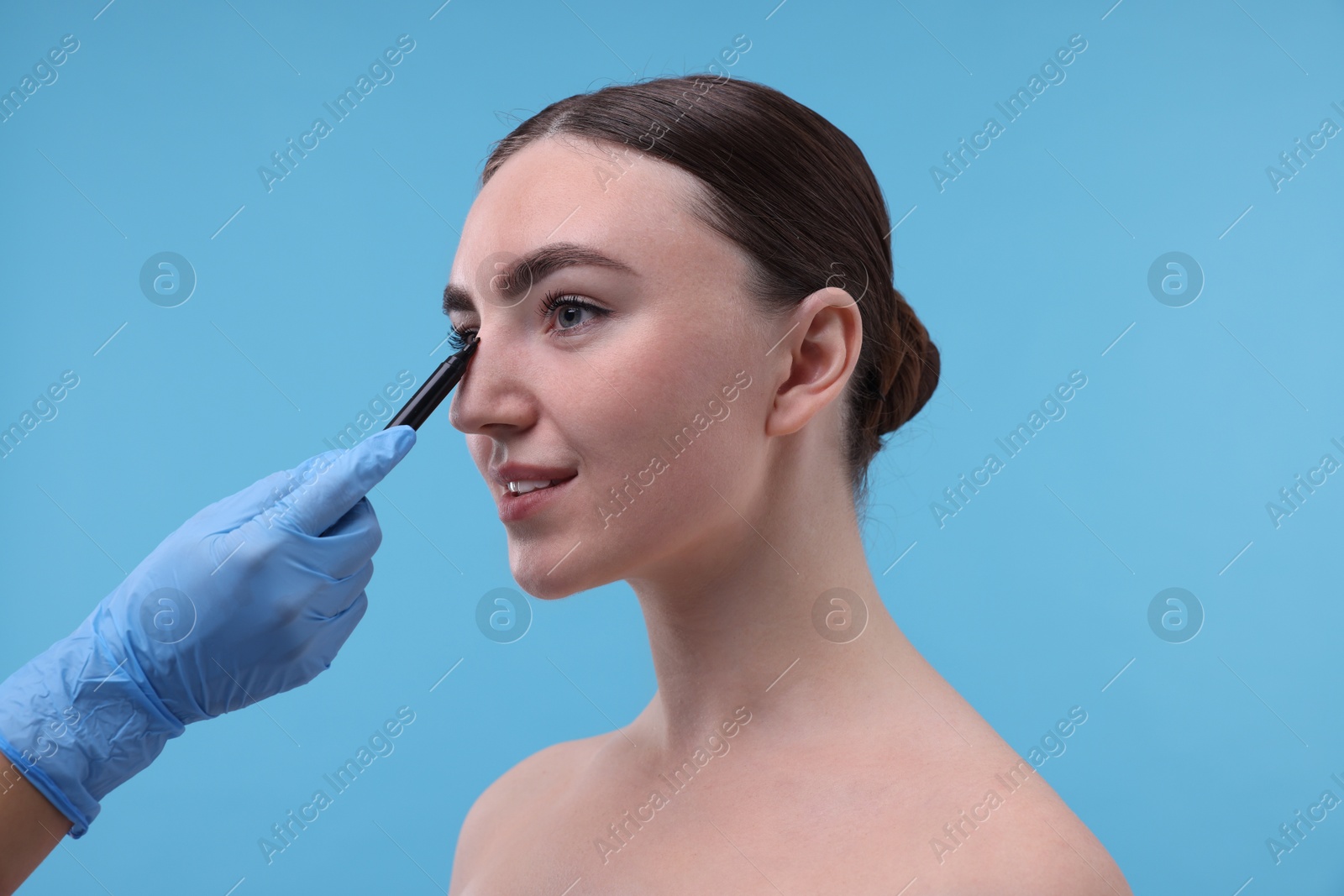 Photo of Doctor drawing marks on patient's nose for plastic surgery operation against light blue background, closeup