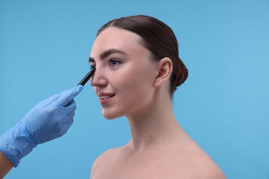 Photo of Doctor drawing marks on patient's nose for plastic surgery operation against light blue background, closeup