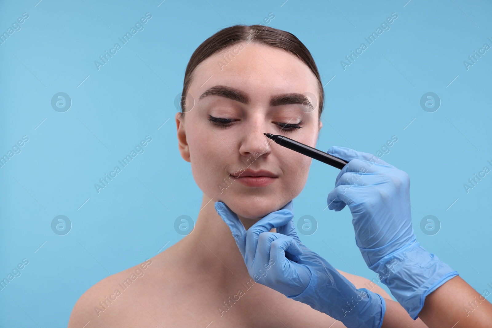 Photo of Doctor drawing marks on patient's nose for plastic surgery operation against light blue background, closeup