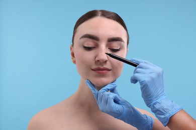 Doctor drawing marks on patient's nose for plastic surgery operation against light blue background, closeup