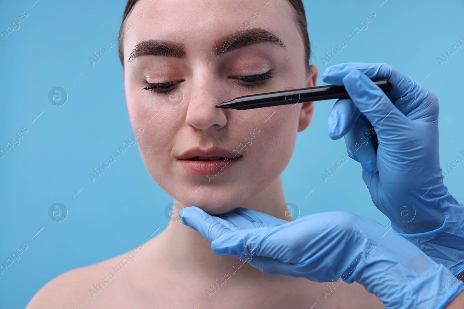 Photo of Doctor drawing marks on patient's nose for plastic surgery operation against light blue background, closeup