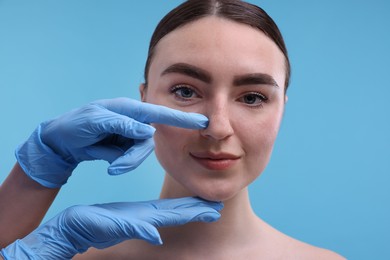 Photo of Doctor checking patient's nose before plastic surgery operation on light blue background, closeup