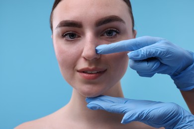 Photo of Doctor checking patient's nose before plastic surgery operation on light blue background, closeup