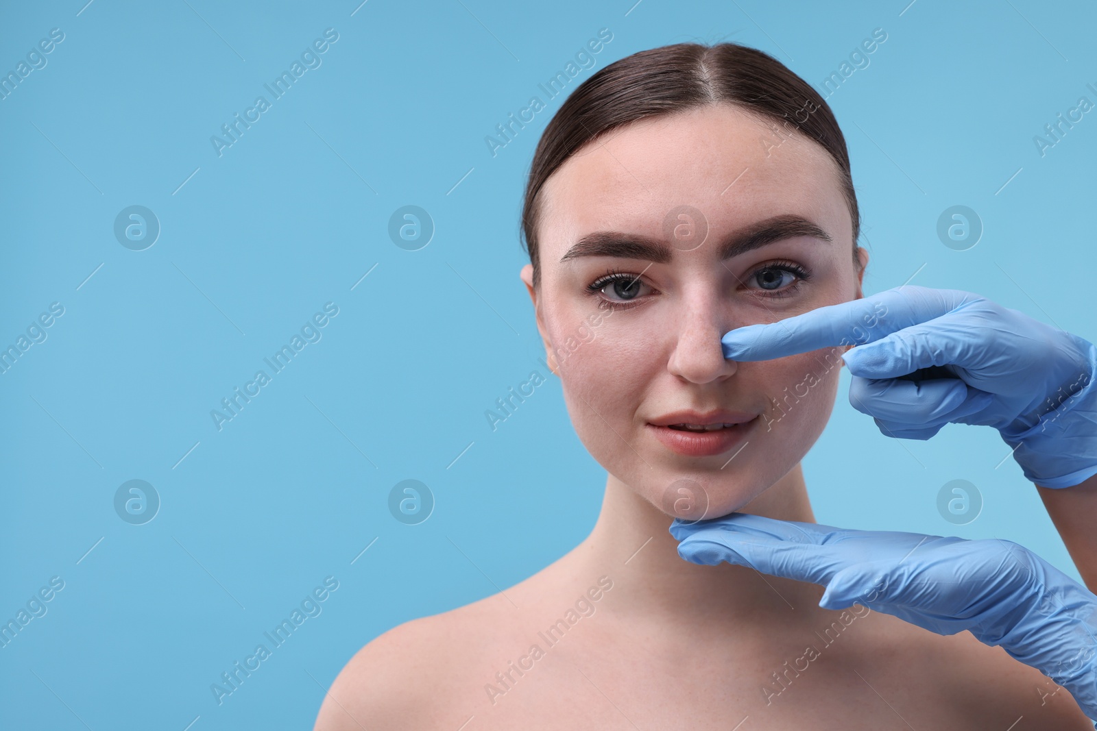 Photo of Doctor checking patient's nose before plastic surgery operation on light blue background, closeup. Space for text