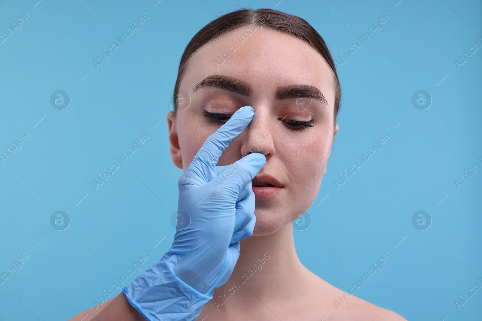 Photo of Doctor checking patient's nose before plastic surgery operation on light blue background, closeup