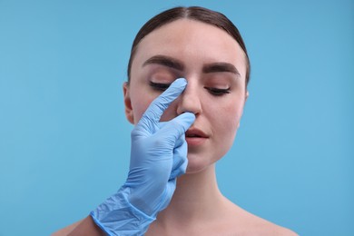 Doctor checking patient's nose before plastic surgery operation on light blue background, closeup