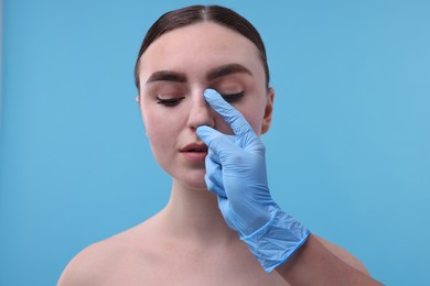 Photo of Doctor checking patient's nose before plastic surgery operation on light blue background, closeup