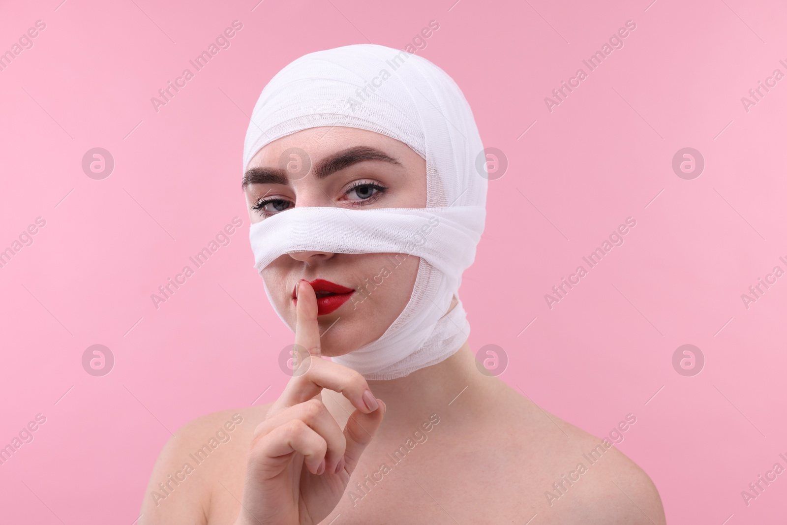 Photo of Woman with nose wrapped in medical bandage after plastic surgery operation making silent gesture on pink background