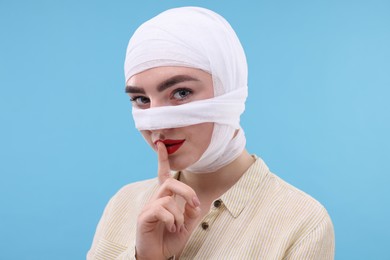 Woman with nose wrapped in medical bandage after plastic surgery operation making silent gesture on light blue background