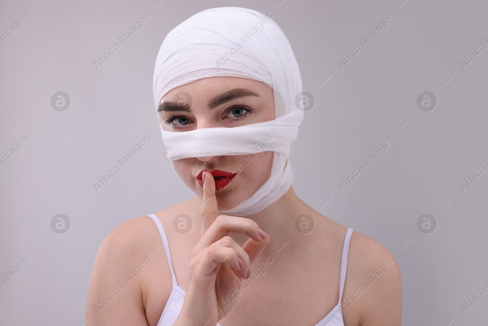 Photo of Woman with nose wrapped in medical bandage after plastic surgery operation making silent gesture on grey background