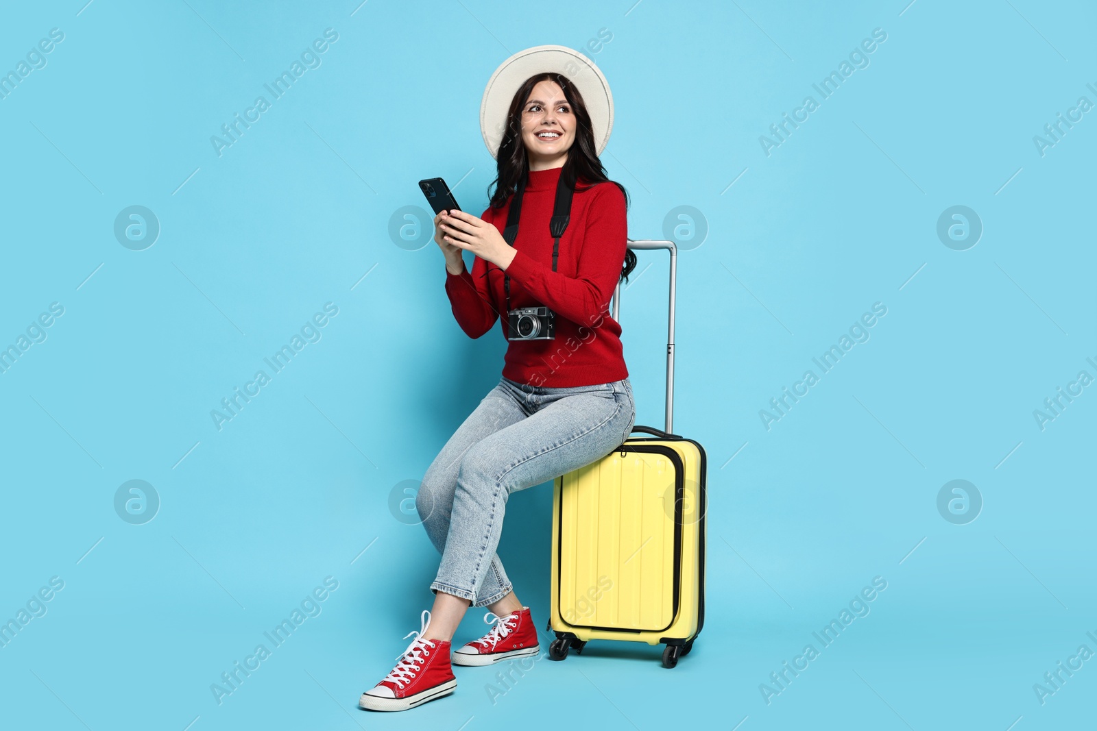 Photo of Young tourist in hat with camera, phone and suitcase on light blue background