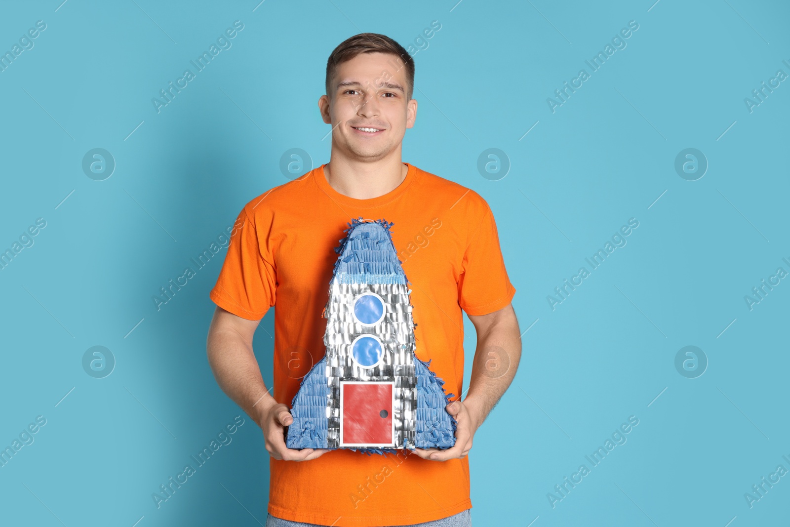 Photo of Happy man with pinata in shape of spaceship on light blue background