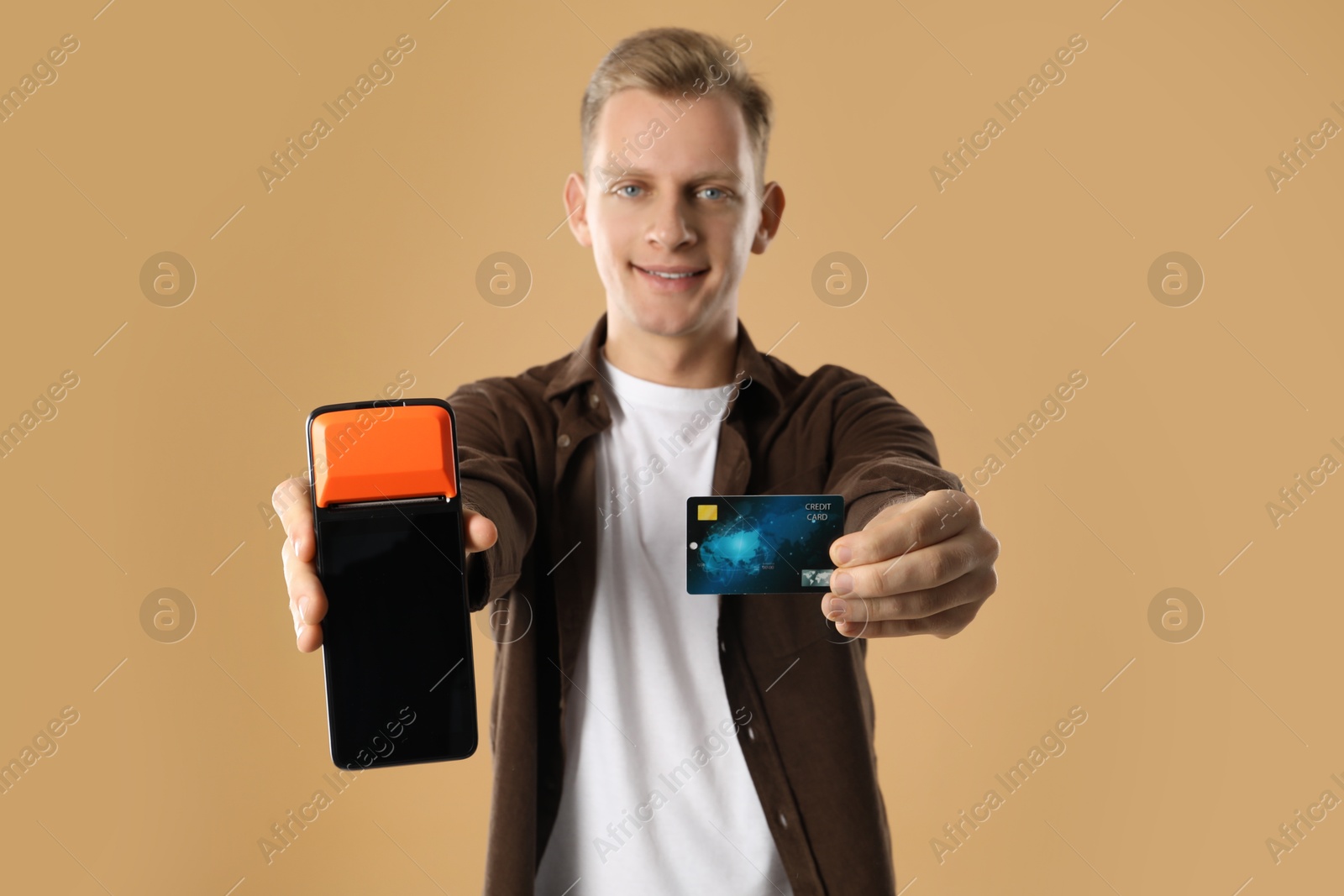 Photo of Happy young man with payment terminal and debit card on beige background