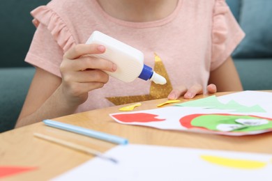 Girl applying glue onto paper figure for her creative project at table indoors, closeup. Art and craft