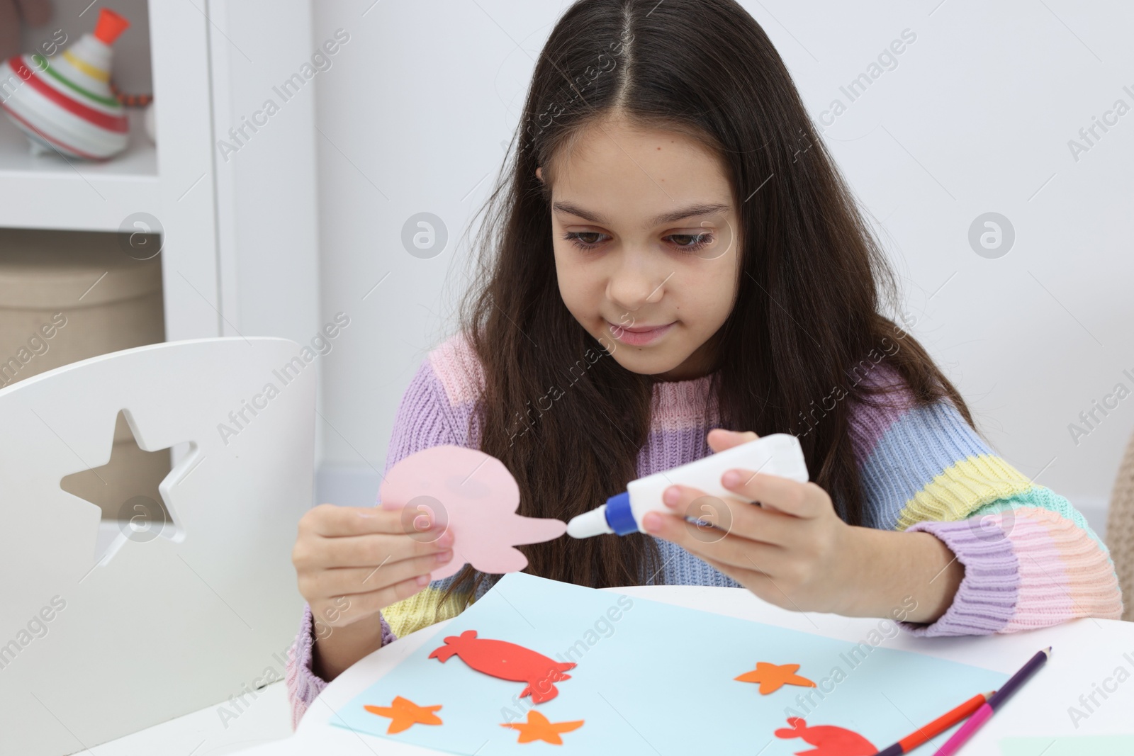 Photo of Girl applying glue onto paper figure for her creative project at table indoors. Art and craft