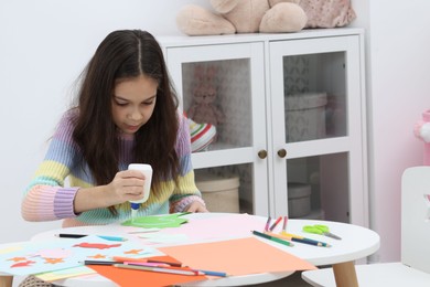 Photo of Girl applying glue onto paper figure for her creative project at table indoors. Art and craft