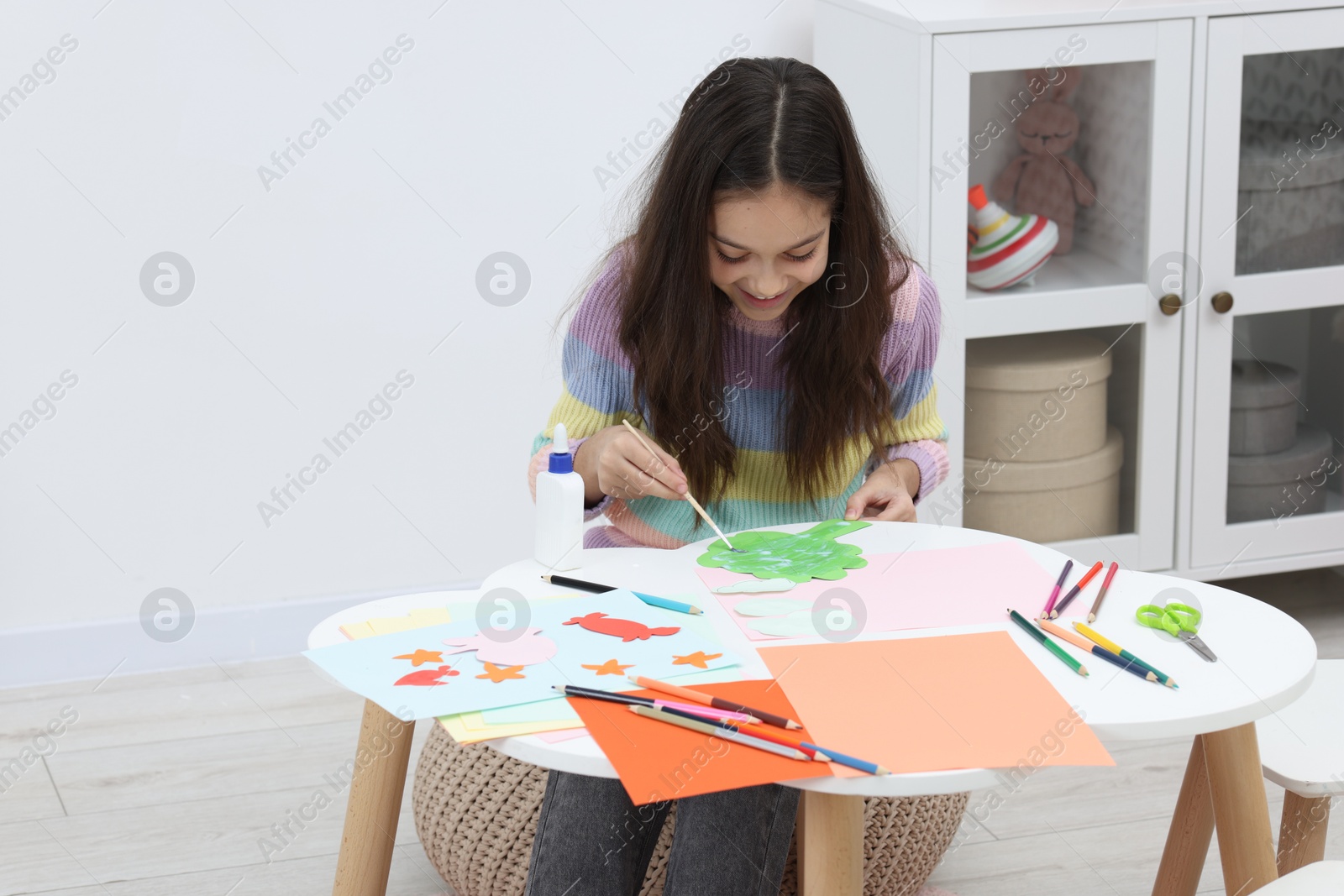 Photo of Girl applying glue onto paper figure for her creative project at table indoors. Art and craft