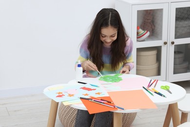 Photo of Girl applying glue onto paper figure for her creative project at table indoors. Art and craft