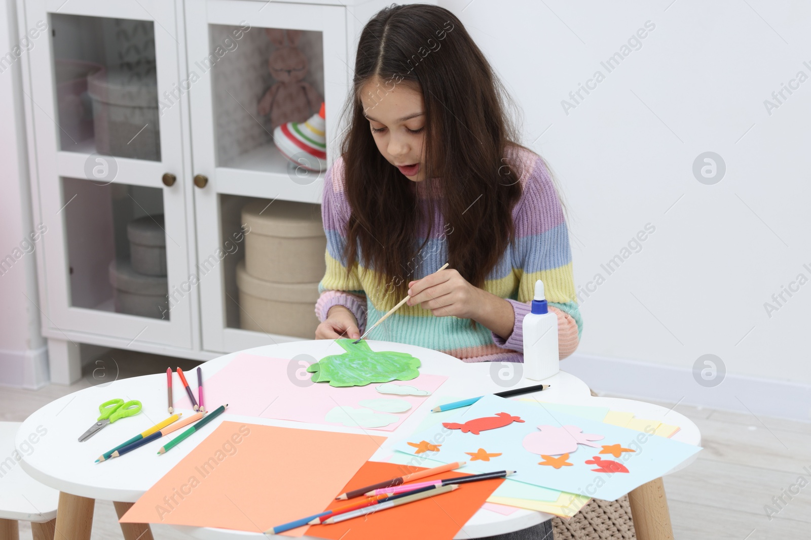 Photo of Girl applying glue onto paper figure for her creative project at table indoors. Art and craft