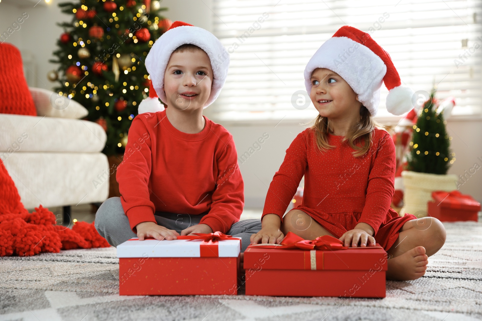 Photo of Happy little children in Santa hats with Christmas gifts on floor at home