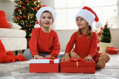 Photo of Happy little children in Santa hats with Christmas gifts on floor at home