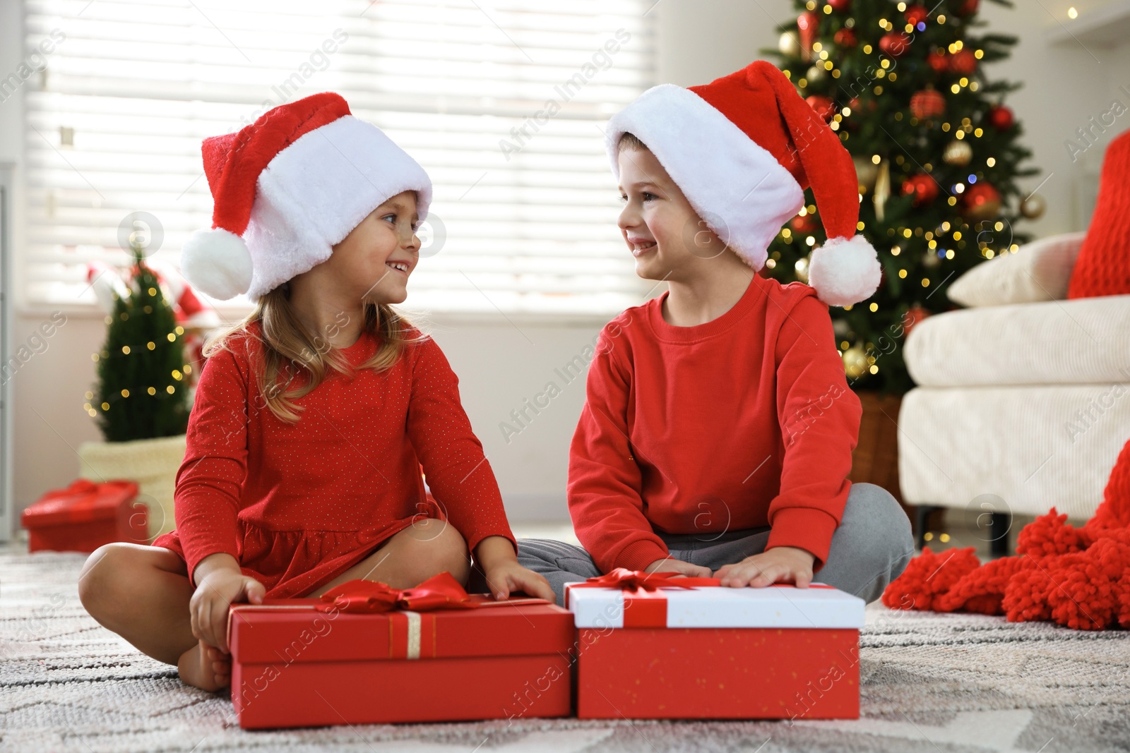 Photo of Happy little children in Santa hats with Christmas gifts on floor at home