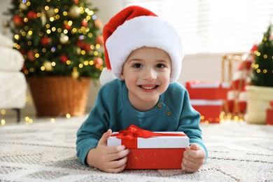 Photo of Happy little boy in Santa hat with Christmas gift on floor at home