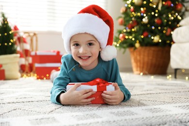 Photo of Happy little boy in Santa hat with Christmas gift on floor at home