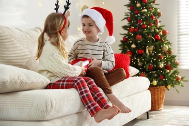 Happy little boy in Santa hat presenting his sister with Christmas gift on sofa at home