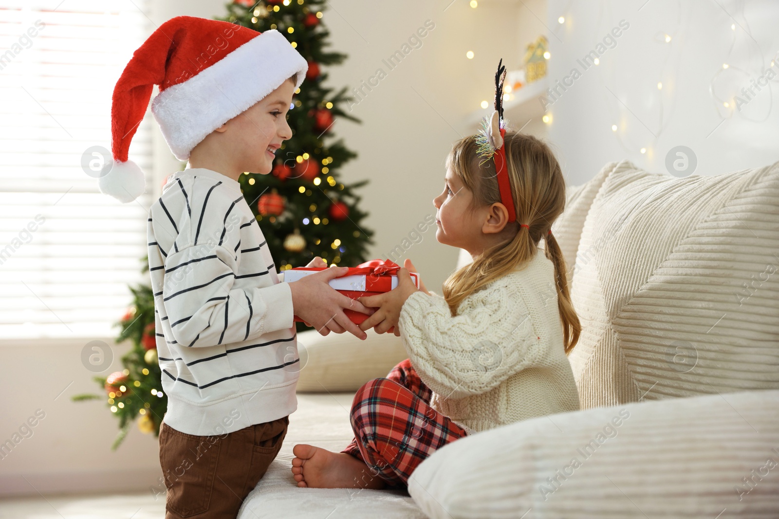 Photo of Happy little boy in Santa hat presenting his sister with Christmas gift at home