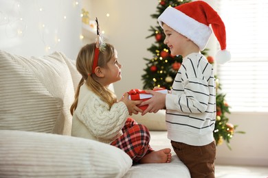 Photo of Happy little boy in Santa hat presenting his sister with Christmas gift at home