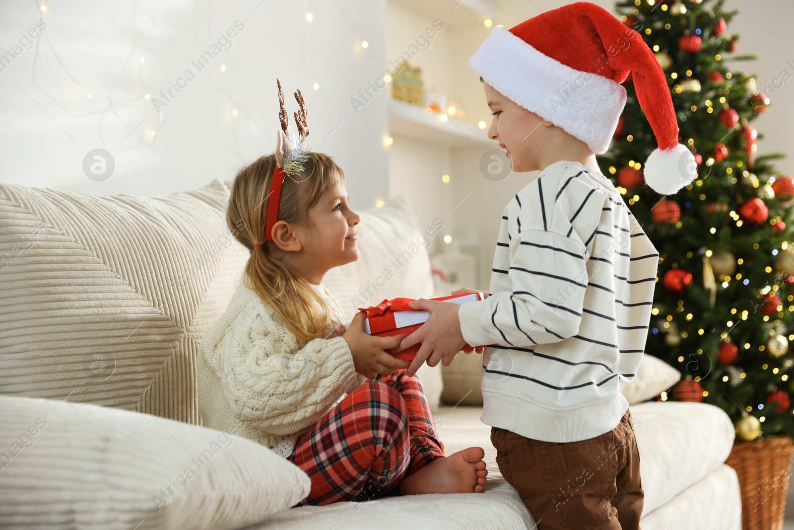 Photo of Happy little boy in Santa hat presenting his sister with Christmas gift at home