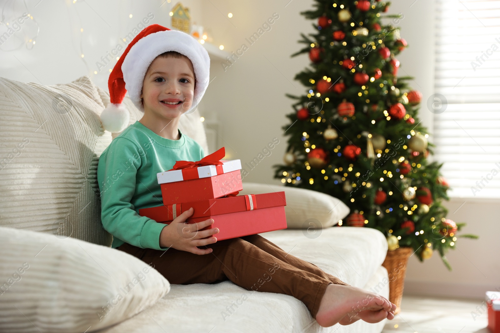 Photo of Happy little boy in Santa hat with Christmas gifts on sofa at home