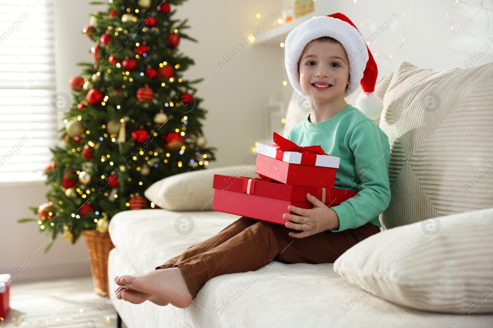 Photo of Happy little boy in Santa hat with Christmas gifts on sofa at home