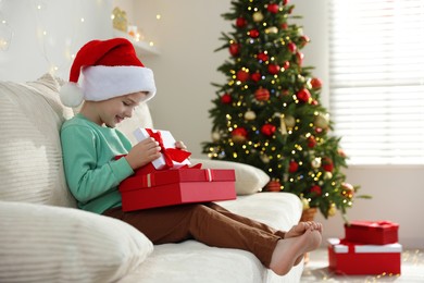 Photo of Happy little boy in Santa hat with Christmas gifts on sofa at home