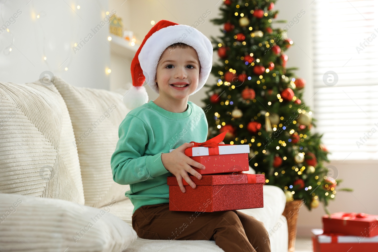 Photo of Happy little boy in Santa hat with Christmas gifts on sofa at home