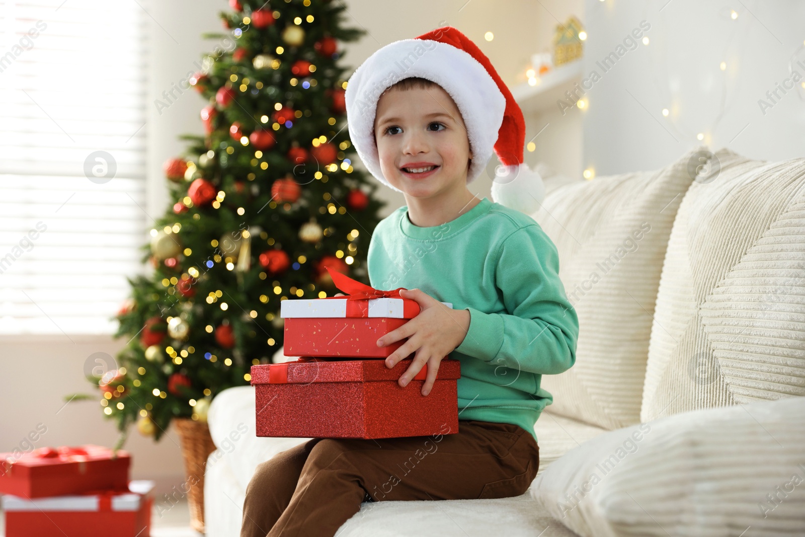 Photo of Happy little boy in Santa hat with Christmas gifts on sofa at home