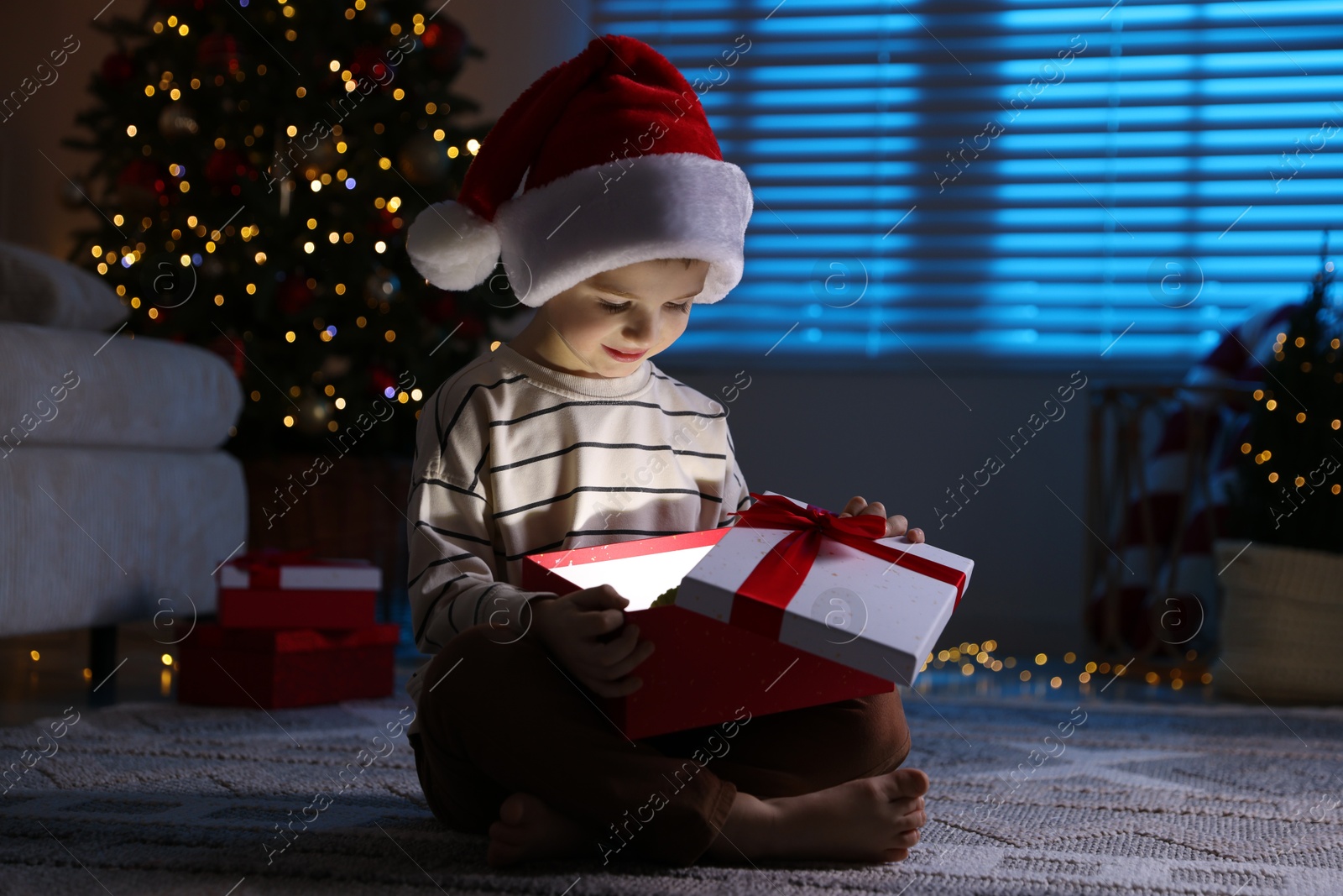 Photo of Little boy in Santa hat with Christmas gift on floor at home